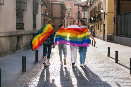 Foto de Vista trasera de cuatro amigos gays, radiante alegría, caminando en una calle soleada de la ciudad al atardecer, adornada con accesorios LGBT, incluyendo banderas, bolsos y camisas. Dos hombres en el centro comparten un beso apasionado. - Imagen libre de derechos
