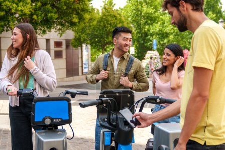 Four cheerful, diverse young people renting electric bikes at a sunny bike rental parking lot in a city. Asian guy arrives with Caucasian girl.