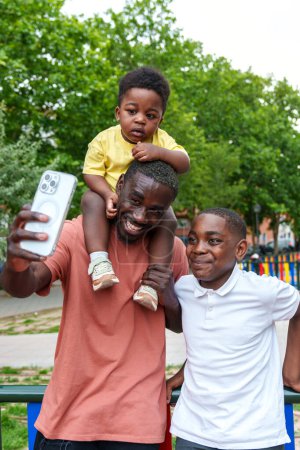 Téléchargez les photos : Rayonnant papa africain claque un selfie vibrant avec ses enfants dans un parc vert coloré ; bébé perché sur les épaules, pur bonheur." - en image libre de droit