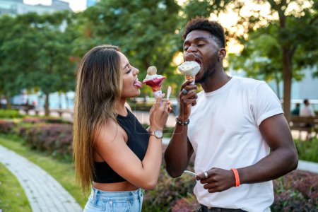 A joyful urban portrait featuring a stylish Afro-American man and a Latina woman sharing a cone of ice cream, surrounded by lush trees and flowers in the warm sunset light.