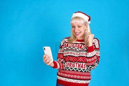Téléchargez les photos : Une femme caucasienne extatique, revêtue de vêtements de Noël rouges, lève le poing dans la joie dans un décor de studio bleu clair, obtenant avec succès son cadeau désiré. Elle adore le Père Noël et Papa Noel. - en image libre de droit