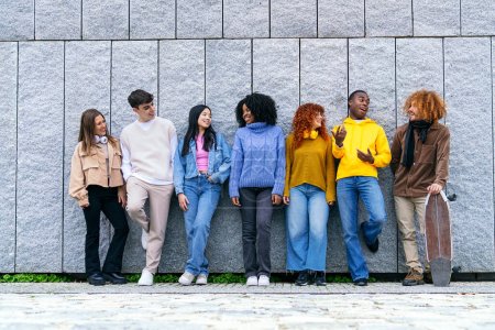 Téléchargez les photos : Groupe diversifié de sept jeunes amis profitant du temps ensemble par un mur gris, mettant en valeur la diversité raciale et l'interaction joyeuse. - en image libre de droit