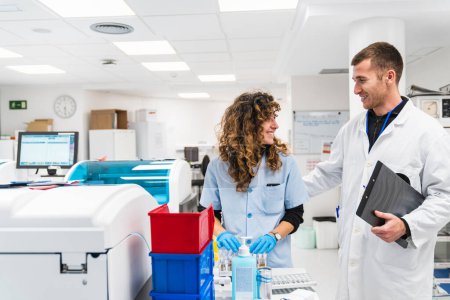 Photo for Two healthcare workers engage in cheerful discussion amidst blood analysis machinery in a contemporary clinical lab setting. - Royalty Free Image