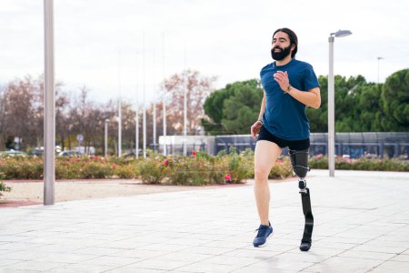 Foto de Alegre corredor barbudo con avanzada pierna protésica de fibra de carbono disfrutando de una carrera en un parque escénico con un cielo nublado telón de fondo - Imagen libre de derechos