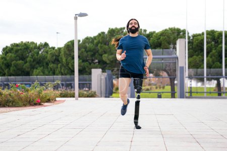Foto de Foto frontal de un hombre barbudo sonriente con una prótesis deportiva de fibra de carbono de última generación, corriendo en un parque urbano. - Imagen libre de derechos
