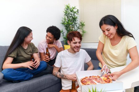 Photo for Multicultural group of friends sitting together on a sofa, enjoying pizza and beers, celebrating a birthday in a warm home setting - Royalty Free Image