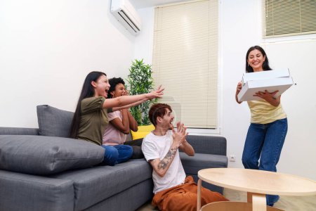 Photo for Multicultural friends excitedly share a large pizza. Happy woman presents the meal to others in a bright, cozy apartment setting - Royalty Free Image