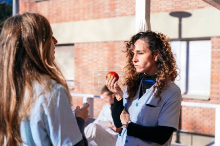 Eine fokussierte Krankenschwester mit Stethoskop genießt einen gesunden Apfel und macht inmitten einer lebhaften Diskussion mit einem Kollegen im Freien eine Denkpause..