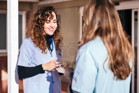 Zwei Krankenschwestern in blauem Peeling genießen ein unbeschwertes Gespräch und eine nahrhafte Mahlzeit in ihrer Pause außerhalb der medizinischen Einrichtung.