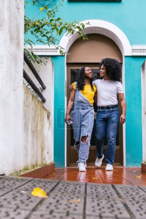 Latinx-Gefährten, einer mit einem großen Afro, bei einem freundlichen Plausch vor einem blauen Hauseingang.