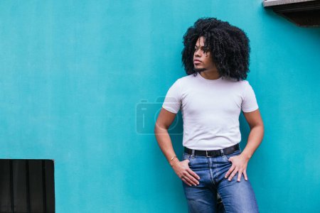Photo for Contemplative Afro-Latino man standing with hands in pockets against a serene turquoise wall, exuding a relaxed urban vibe. - Royalty Free Image