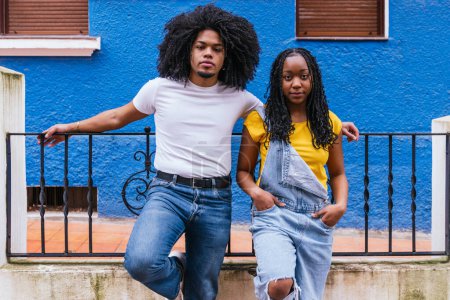 Junges Afro-Latino-Paar in lässiger Kleidung posiert mit ernstem Blick vor einer lebendigen blauen Wand und präsentiert urbanen Stil und Selbstbewusstsein.
