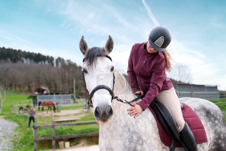 Reiterin in Reiterkleidung feilt am Zaumzeug ihres Schimmels auf einem eingezäunten Außenreitplatz auf einem Bauernhof.