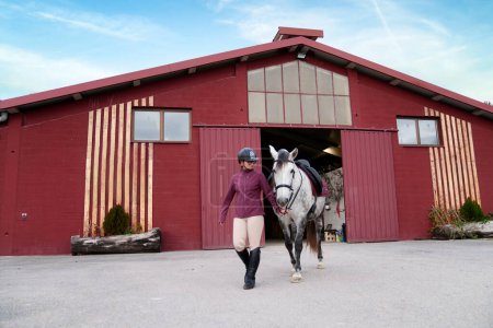 Equestrian in maroon jacket leading her dappled grey horse from an open red barn into the daylight.