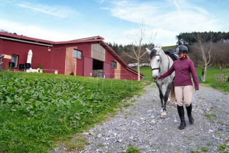 Young rider in equestrian gear walking her grey horse on a farm path beside a red barn and green field.