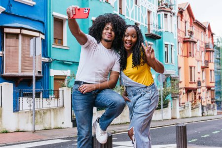 Latino friends posing for a lively selfie on a city street, surrounded by colorful houses, radiating happiness and style.