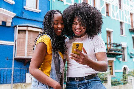 Two Latino friends enjoying a fun moment with a smartphone on a vivid blue balcony, embodying joy and connection.