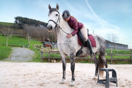 Téléchargez les photos : Jeune cavalier concentré montant un cheval blanc moucheté dans une arène sablonneuse ouverte avant de commencer la pratique équestre - en image libre de droit