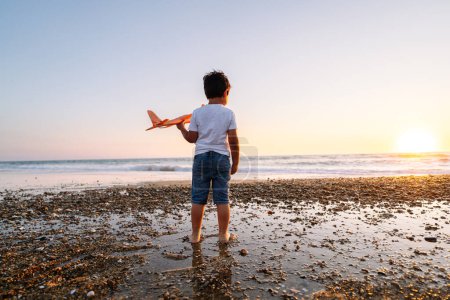 Niño retroiluminado con avión en un paisaje marino al atardecer.