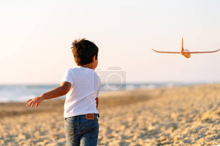 Garçon avec un avion jouet chassant le vent le long du rivage au coucher du soleil.