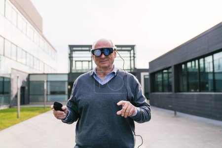 Un hombre de negocios senior que usa gafas de realidad virtual y usa un controlador, parado fuera de un moderno edificio de oficinas.