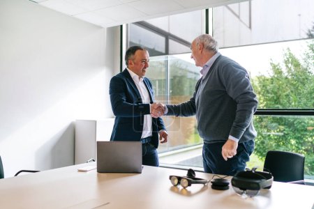 Two men, a businessman and a client, shake hands after a successful business deal on VR glasses in a modern office environment.