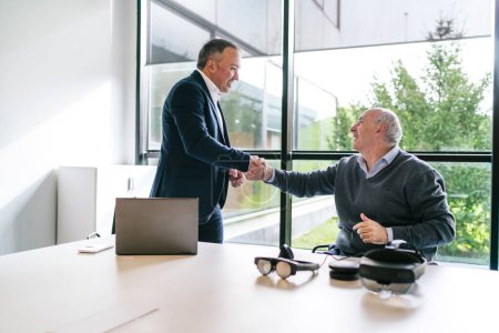 A businessman and client shake hands after finalizing a deal for VR glasses in a modern, bright office setting.