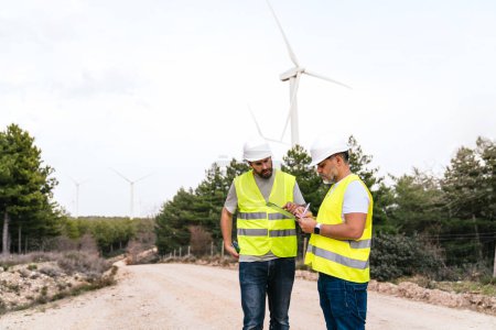 Two engineers in safety gear discuss wind turbine maintenance. They stand on a dirt road, surrounded by trees and wind turbines.