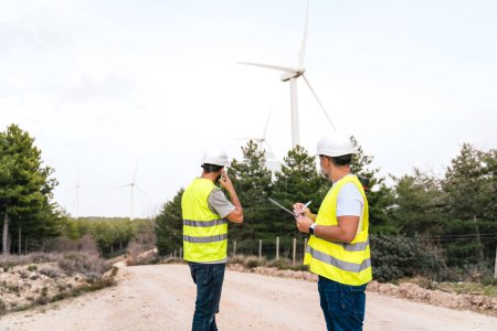 Two engineers, one using a phone, the other writing on a clipboard, examine wind turbines. The scene is set on a dirt road with a forest background.