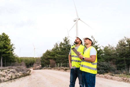 Two engineers in high-visibility vests inspect wind turbines. They stand on a dirt road with a forest and turbines in the background.