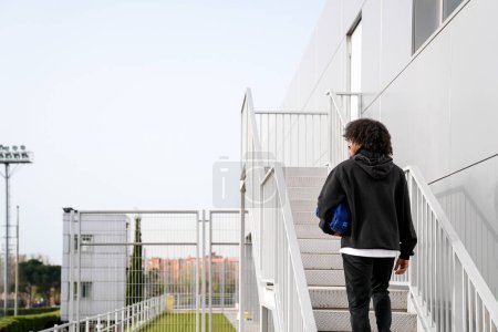 A African person wearing a black hoodie is walking up a set of white stairs. The person is carrying a blue bag. The scene is at the entrance of a sports center