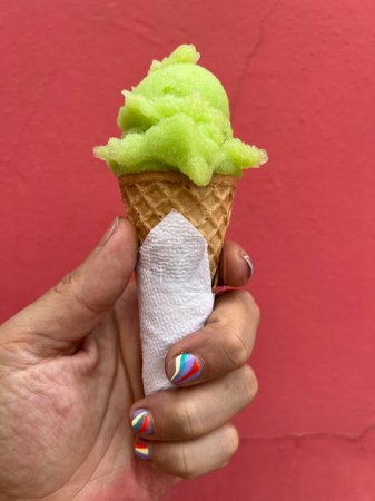 Foto de La mano del hombre sostiene una bola verde de helado de lima en un cono contra una pared roja. El hombre tiene una manicura de uñas. - Imagen libre de derechos