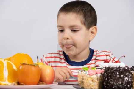 Un garçon d "âge préscolaire choisit ce qu'il faut manger - des fruits ou des gâteaux, choisit des fruits et lèche.