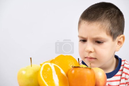 Foto de Un niño en edad preescolar mira las frutas - naranjas, caquis y manzanas sobre un fondo blanco, un lugar para el texto. - Imagen libre de derechos