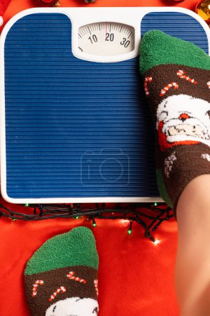 Foto de Una mujer en calcetines de Navidad se para en la balanza, se pesa después de la gula durante las vacaciones. - Imagen libre de derechos