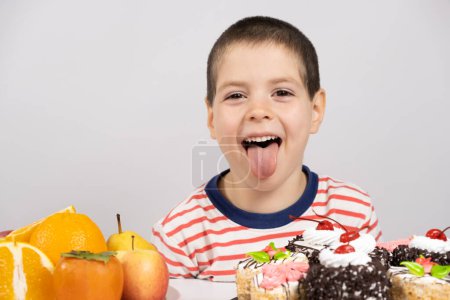5 ans mignon garçon assis devant des fruits et des gâteaux et choisit ce qu'il faut manger.