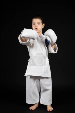 Téléchargez les photos : Un petit garçon apprend les arts martiaux, fait du karaté en kimono et des gants de boxe. - en image libre de droit