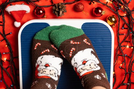 Téléchargez les photos : Une femme en chaussettes de Noël se tient sur la balance, se pèse après la gourmandise pendant les vacances. - en image libre de droit