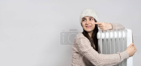 Photo for A woman in a hat hugs an oil heater and points her finger to the side in place for text - Royalty Free Image