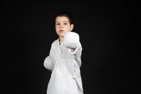 Téléchargez les photos : Un petit garçon apprend les arts martiaux, fait du karaté en kimono et des gants de boxe. - en image libre de droit