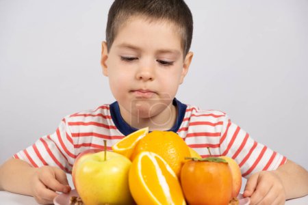 Téléchargez les photos : Un garçon d "âge préscolaire regarde les fruits - oranges, kaki et pommes sur fond blanc - en image libre de droit