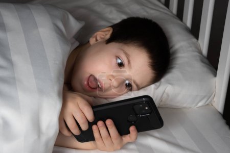 Photo for A 5 year old boy plays games on his phone while lying in bed. Gadgets in bed before bedtime in a child. - Royalty Free Image