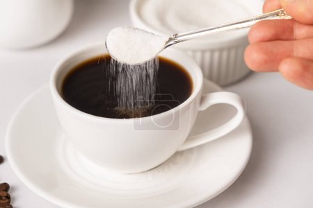 Téléchargez les photos : Le processus de verser le sucre blanc d'une cuillère dans une tasse de café blanc. Accro au sucre, diabète - en image libre de droit