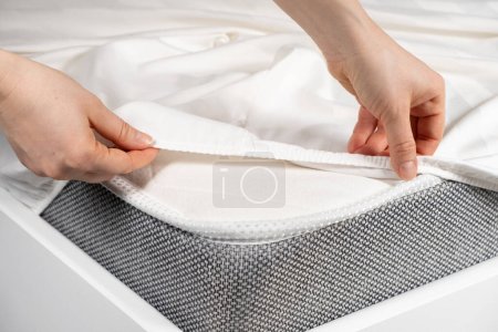 Foto de Una sábana de satén blanco con una banda elástica se pone en el colchón de la cama, un primer plano de las manos. Ropa de cama - Imagen libre de derechos