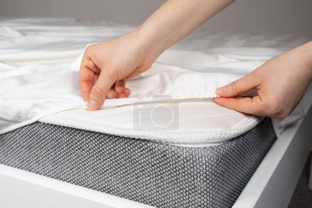 Foto de Una sábana de satén blanco con una banda elástica se pone en el colchón de la cama, un primer plano de las manos. Ropa de cama - Imagen libre de derechos