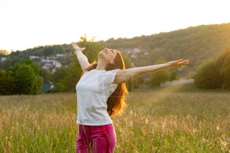 Eine Frau steht mit erhobenen Händen in der untergehenden Sonne im Sommer auf einem Feld in der Natur.