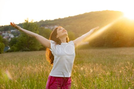 A woman stands in a field in nature with her hands raised in the rays of the setting sun in summer