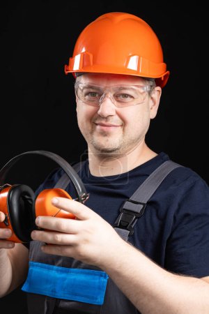 Téléchargez les photos : Bâtisseur dans un casque de protection avec des écouteurs anti-bruit dans ses mains sur un fond noir. - en image libre de droit