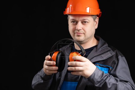 Foto de Constructor en un casco protector con auriculares que cancelan el ruido en sus manos sobre un fondo negro, copiar espacio para el texto. - Imagen libre de derechos