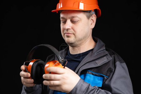 Foto de Constructor en un casco protector con auriculares que cancelan el ruido en sus manos sobre un fondo negro. - Imagen libre de derechos